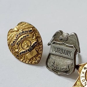 Puede incluir: Tres insignias de metal dorado. La primera insignia es de forma de escudo con el texto "California Plant Protection". La segunda insignia es de forma de escudo con el texto "Burbank Police". La tercera insignia es de forma de estrella de seis puntas con el texto "Kern County Sheriff".