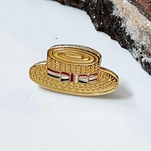 May include: A gold-coloured hat pin in the shape of a straw hat. The hat has a red, white, and blue striped band. The pin is likely made of metal and has a detailed, textured surface.