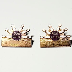 May include: Two gold-colored 500 Club bowling pins lapel pins. Each pin features a white circle with the text "500 CLUB" in black lettering. The circle is surrounded by a ring of white bowling pins.