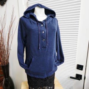 Puede incluir: Una sudadera con capucha azul marino con un bolsillo delantero y cierre de tres botones. La parte superior de manga larga tiene un cordón y se muestra en un maniquí. La prenda está hecha de un material suave.