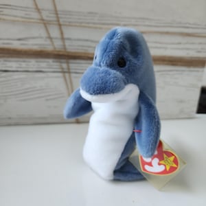 Puede incluir: Un peluche de delfín azul con vientre blanco y etiqueta adjunta. El delfín tiene ojos negros y está hecho de material suave. La etiqueta es roja y amarilla.