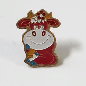 May include: A vintage enamel pin featuring a smiling cartoon cow with a red body, white face, and a flower on its head. The pin has gold-colored trim and is set against a white background. The cow is holding a blue and yellow object.