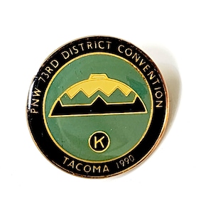 Könnte beinhalten: Runder Ansteckpin mit dem Text "PNW 73RD DISTRICT CONVENTION TACOMA 1990". Der Pin hat eine grüne Mitte mit einem goldenen und schwarzen Bergdesign. Der äußere Ring ist schwarz mit goldfarbenen Buchstaben.