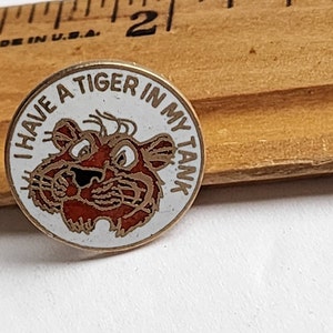 May include: A round metal pin with a white background and a gold-tone border. The pin features a cartoon tiger face in brown and black, with the text "I HAVE A TIGER IN MY TANK" arched above it. A wooden ruler is visible, showing the pin is 1 inch wide.