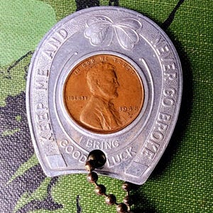 May include: A silver horseshoe-shaped charm with a 1948 penny at its center. The charm has the words "KEEP ME AND NEVER GO BROKE" and "BRING GOOD LUCK" engraved on it. A small chain is attached.