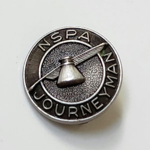 May include: A circular, silver-toned pin with the text "NSPA JOURNEYMAN" around the edge. The center features a stylized anvil with wings. The pin has a textured background and a detailed design.