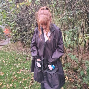 May include: A dark gray raincoat with a collar and pockets. The pockets are decorated with heart and leaf-shaped appliqués. A blue bow is attached to the heart appliqué. The person is wearing dark jeans and sneakers.