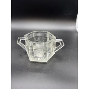 May include: A clear, faceted glass sugar bowl with two handles. The bowl has a hexagonal shape with etched floral designs on the front and sides. The glass has a ribbed texture and is set against a dark background.