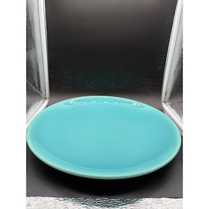 May include: A round, solid turquoise ceramic plate with a smooth, glossy finish and a slightly raised rim. The plate features a simple, minimalist design with a uniform color throughout, showcasing a clean, circular shape and a reflective surface.