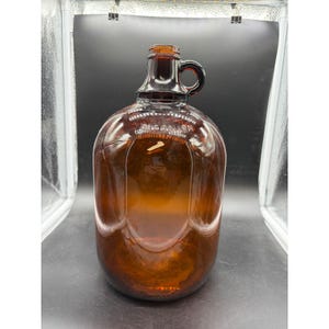 May include: A brown glass jug with a handle, likely for beverages. The jug has a rounded body and a narrow neck with a small handle. The text "1/2 GALLON" is visible on the jug. The glass has a warm, amber hue.