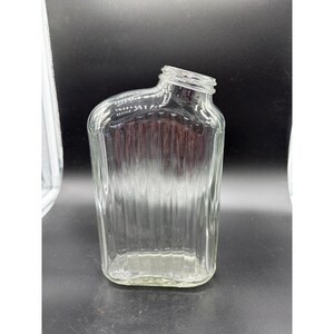 May include: A clear, rectangular glass bottle with a slanted neck and a ridged design. The bottle is empty and sits upright against a dark background. The bottle's shape is reminiscent of a flask.