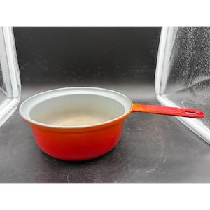 May include: A small, round, red saucepan with a long handle. The interior is a light gray color, and the rim is white. The handle transitions from orange to red. This vintage cookware is ideal for cooking small portions.