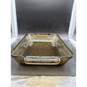 May include: A rectangular baking dish made of translucent amber-colored glass. The dish features integrated handles on two opposite sides and a smooth, polished rim. The base of the interior displays embossed text indicating the brand and dimensions.