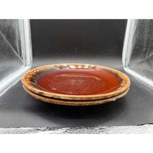 May include: Two stacked ceramic plates. The plates have a brown center and a speckled, tan rim. The plates are a warm, earthy tone, and the glossy finish reflects light. The plates are likely for serving food.