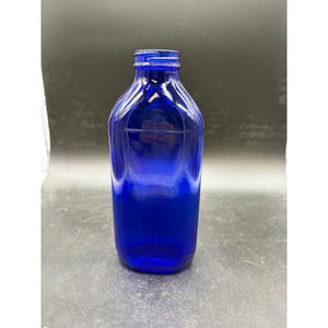 May include: A cobalt blue glass bottle with a rectangular body and rounded corners. The bottle features a narrow neck with a threaded top finish, designed for a screw-on cap. The glass is translucent and smooth, reflecting light on its surface.
