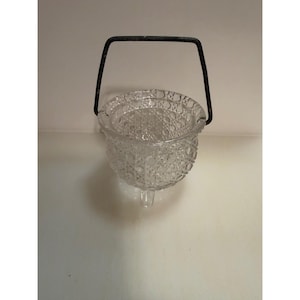 May include: A clear glass bucket with a textured diamond and circle pattern on the exterior. The bucket features a dark metal swing handle attached to the rim and stands on three small, clear glass feet at the base of the vessel.