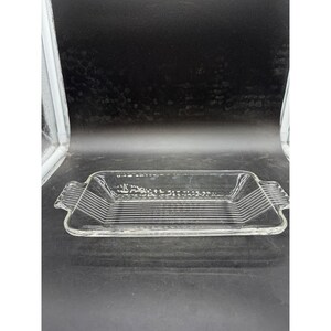 May include: A clear rectangular glass baking dish featuring integrated side handles and a series of parallel horizontal lines embossed along the interior base and extending up the sides, providing a textured appearance to the transparent glass.