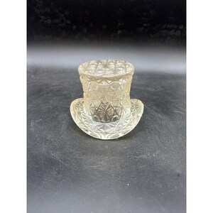 May include: A clear, colorless glass top hat figurine featuring a pressed diamond and starburst geometric pattern across the entire surface. The object has a cylindrical crown and a curved brim, with a textured, faceted finish throughout the glass.