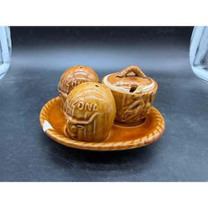 May include: A vintage ceramic condiment set in shades of brown. The set includes two salt and pepper shakers and a lidded sugar bowl with a spoon, all resting on a matching plate. The word "Arizona" is embossed on the shakers.