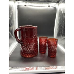 May include: A ruby red glass pitcher and two matching glasses. The pitcher has a large handle and a textured surface with small, raised bumps. The glasses have a similar design. The set is likely for serving beverages.