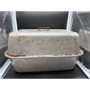 May include: A rectangular, vintage aluminum roasting pan with a lid. The pan has a handle on the lid and shows signs of wear, including discoloration and rust spots. The pan is designed for cooking and baking.
