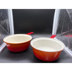 May include: Two orange enameled cast iron saucepans with white interiors and red handles. The pans are oval-shaped and appear unused. The handles are short and angled. The background is a studio setup with a black backdrop.