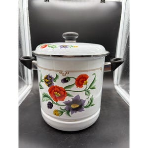 May include: A white enamel stockpot with a matching lid, featuring a colorful floral pattern of red, purple, and yellow flowers with green leaves. The pot has two black side handles and a black knob on the lid, with a thin gold decorative line.