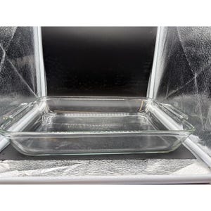 May include: A clear, square glass baking dish with integrated handles on opposite sides. The dish features a smooth, transparent surface and a flat bottom, designed for oven use. The glass is thick and durable, with rounded corners and edges.