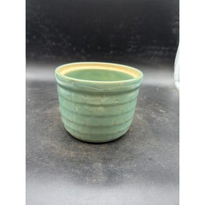 May include: A sage green ceramic bowl with a textured, grid-like pattern on the exterior. The bowl features a smooth, rounded rim and a light beige interior. The vessel has a cylindrical shape with slightly tapered sides and a flat base.