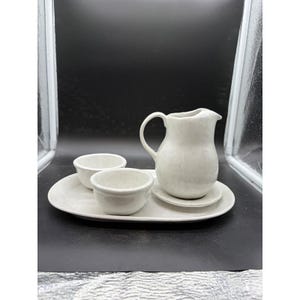 May include: A white ceramic set consisting of an oval serving tray, a small pitcher with a curved handle and spout, and two matching small bowls. The pieces feature a smooth, glossy finish with subtle speckled details throughout the surface.
