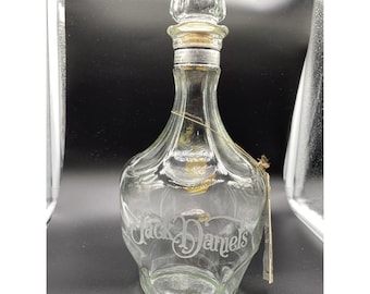 Könnte beinhalten: Transparente Jack Daniel's Glas-Dekantierkaraffe mit dekorativem Verschluss. Die Karaffe hat einen schlanken Hals und einen bauchigen Körper. Die Worte "Jack Daniel's" sind auf der Vorderseite eingraviert. Ein Korken und ein goldfarbenes Metallband verschließen die Flasche.
