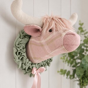 May include: A plush Highland cow head wall decor. The cow head is pink plaid with a pink fringe and cream-colored horns. It is surrounded by a green wreath and a pink bow. The decor is mounted on a white wall.