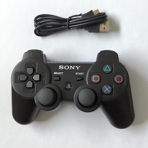 May include: A black Sony PlayStation game controller featuring a directional pad, two analog sticks, and action buttons with triangle, circle, cross, and square symbols. The controller includes Select and Start buttons and comes with a black USB cable.