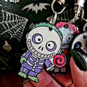 May include: A multi-charm keychain featuring a character with a white skull face, purple and green clothing, and a red and black swirl lollipop. Beside it, a second charm depicts a character with red hair and stitched facial features.