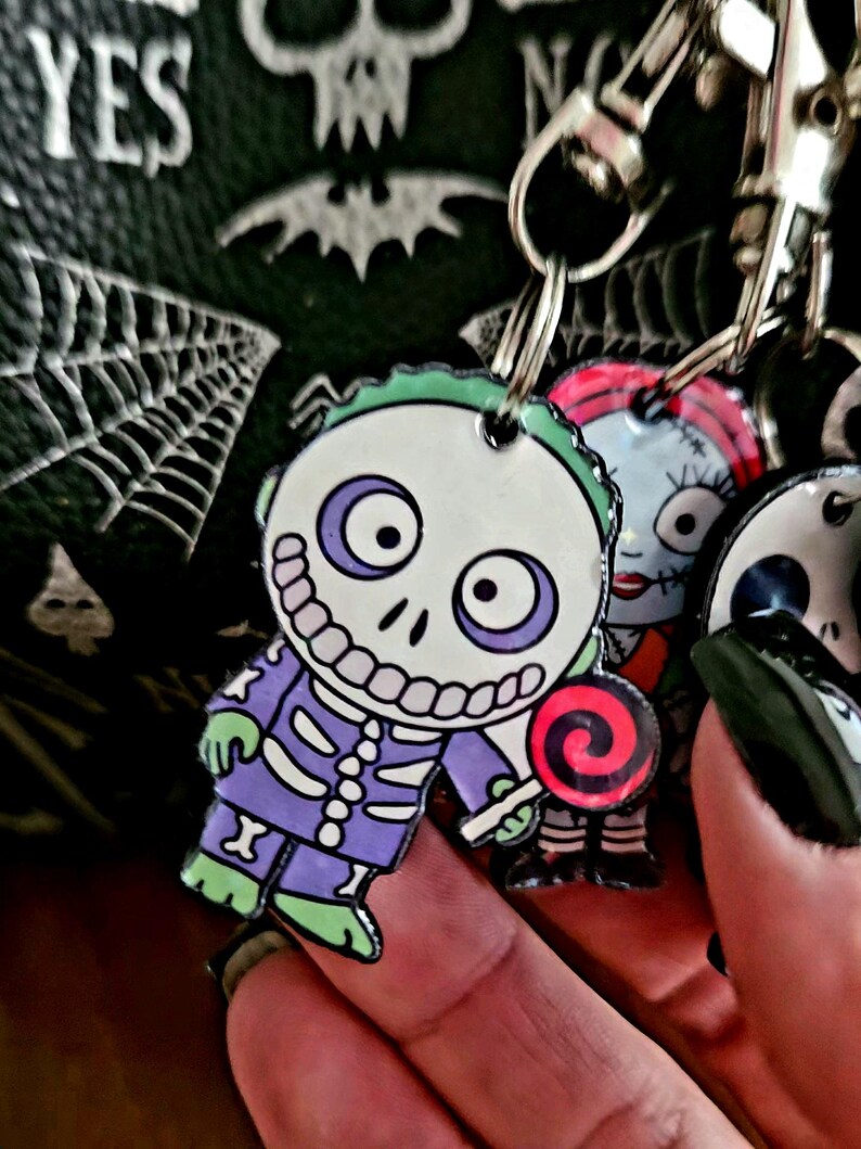 May include: A multi-charm keychain featuring a character with a white skull face, purple and green clothing, and a red and black swirl lollipop. Beside it, a second charm depicts a character with red hair and stitched facial features.