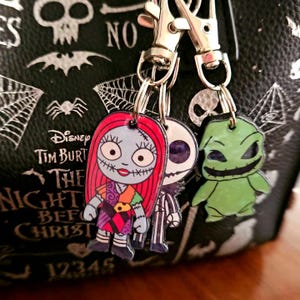May include: A set of three character charms attached to silver-toned metal lobster clasps. The charms feature Sally with red hair and a patchwork dress, Jack Skellington in a pinstripe suit, and a green Oogie Boogie figure with a wide grin.