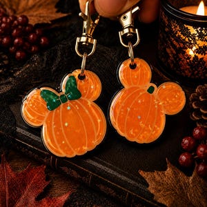 May include: Two orange pumpkin-shaped acrylic keychains with gold-tone metal lobster clasps. One keychain features a dark green bow, and both have a glittery finish with small star-shaped accents embedded within the translucent orange material.