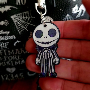 May include: A character keychain featuring a white skull head with dark purple star-filled eyes and a wide stitched smile. The figure wears a black and white pinstriped suit with a bat-shaped bowtie, attached to a silver metal lobster claw clasp.