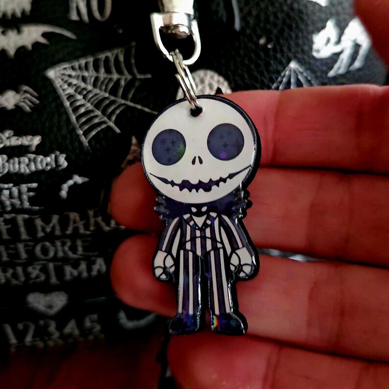 May include: A character keychain featuring a white skull head with dark purple star-filled eyes and a wide stitched smile. The figure wears a black and white pinstriped suit with a bat-shaped bowtie, attached to a silver metal lobster claw clasp.