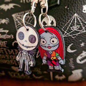 May include: Two character charms featuring Jack Skellington and Sally from The Nightmare Before Christmas. Each charm is attached to a silver-toned metal lobster clasp by a small split ring. The figures are flat, printed designs with detailed facial features and clothing patterns.