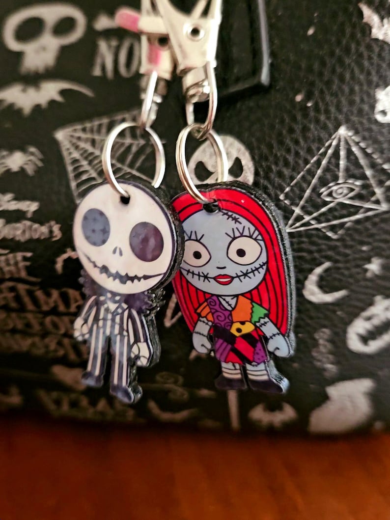 May include: Two character charms featuring Jack Skellington and Sally from The Nightmare Before Christmas. Each charm is attached to a silver-toned metal lobster clasp by a small split ring. The figures are flat, printed designs with detailed facial features and clothing patterns.