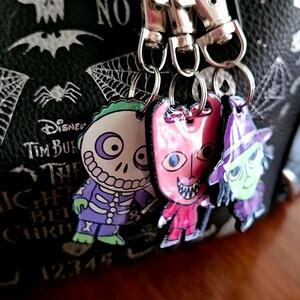 May include: Three character keychains featuring Barrel, Lock, and Shock from The Nightmare Before Christmas, attached to silver metal lobster clasps. The charms are colorful, flat, and hang against a black bag with white skull and spider web prints.
