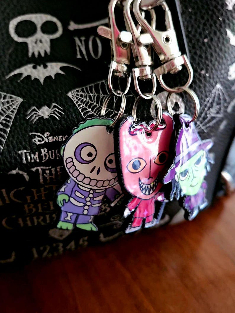 May include: Three character keychains featuring Barrel, Lock, and Shock from The Nightmare Before Christmas, attached to silver metal lobster clasps. The charms are colorful, flat, and hang against a black bag with white skull and spider web prints.