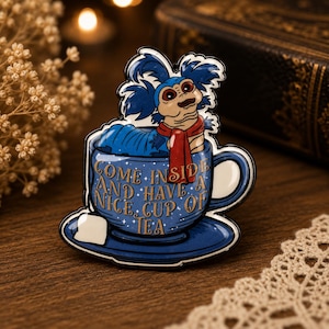 May include: A blue enamel pin featuring a caterpillar with blue hair and a red scarf sitting inside a blue teacup on a saucer. The teacup has gold text that reads, "COME INSIDE AND HAVE A NICE CUP OF TEA," with small white stars and a white sugar cube.