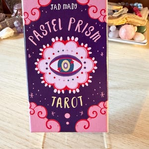 Op de afbeelding: Een rechthoekige doos voor een tarotdeck, getiteld "Pastel Prism Tarot" in witte en roze letters. De doos is paars met een bloemig oogontwerp in het midden. De doos wordt weergegeven op een heldere acrylstandaard.