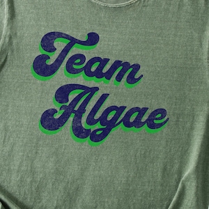 May include: A sage green short-sleeve t-shirt featuring the text Team Algae printed in a dark navy blue script font with a bright green outline. The garment has a crew neckline and a slightly distressed, vintage-inspired fabric texture.