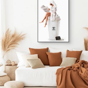 May include: A framed art print depicts a woman in a white robe and towel, reading a newspaper while seated on a large white coffee cup with the Chanel logo. A black purse rests beside the cup. The artwork is displayed above a white sofa with brown and beige pillows.