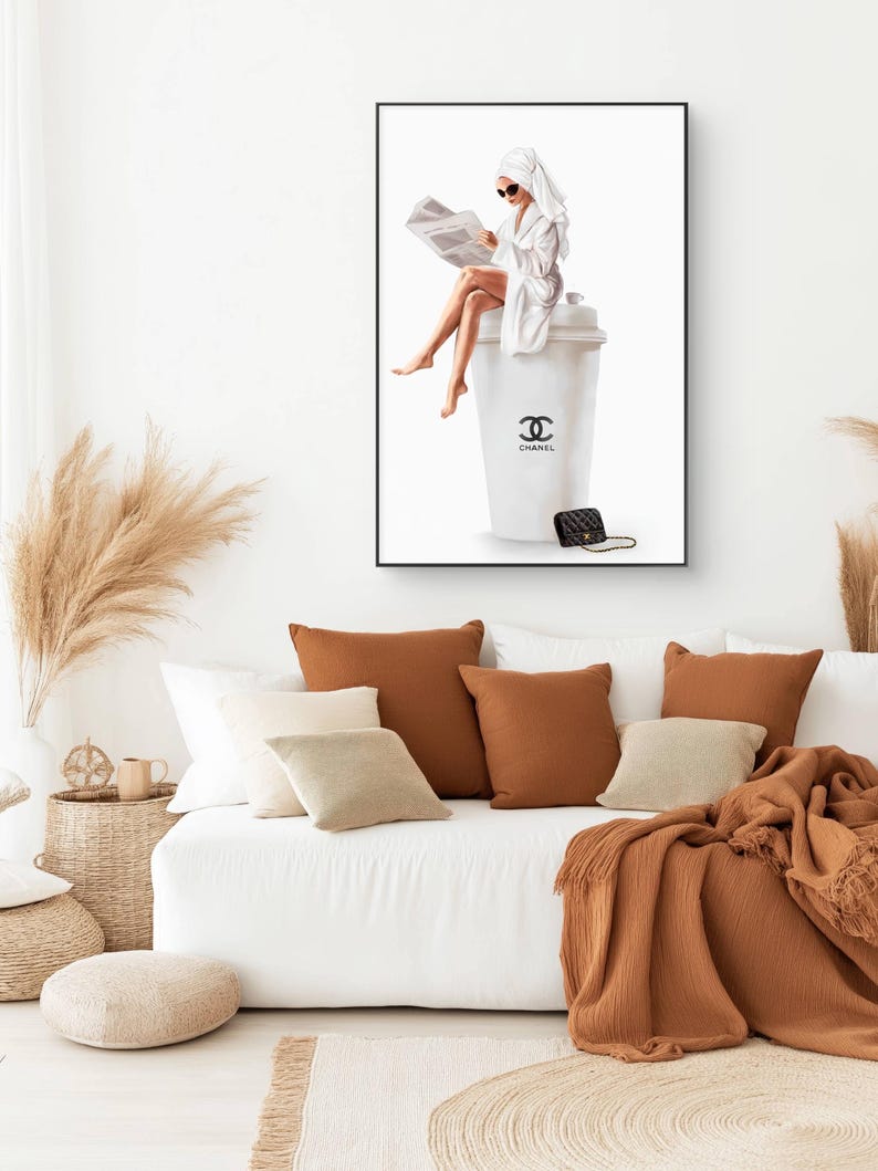 May include: A framed art print depicts a woman in a white robe and towel, reading a newspaper while seated on a large white coffee cup with the Chanel logo. A black purse rests beside the cup. The artwork is displayed above a white sofa with brown and beige pillows.