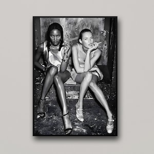 May include: A black and white photographic print featuring two individuals sitting side by side. One wears a metallic sleeveless top and layered necklaces, while the other wears a light-colored tank top. Both are holding cigarettes in their hands.