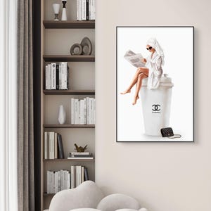 May include: A framed art print depicts a woman in a white robe and towel, reading a newspaper while seated on a large white coffee cup. The cup features the Chanel logo and a small black purse rests nearby. The artwork is displayed in a modern interior.